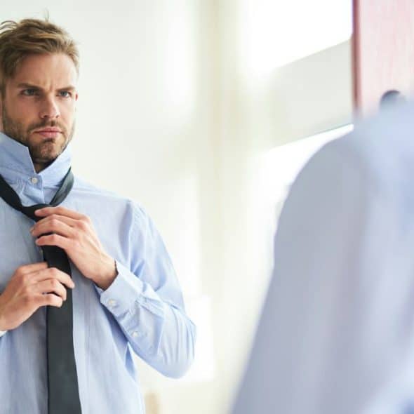 A focused man with a beard is tying a dark tie in front of a mirror while wearing a light blue collared shirt.