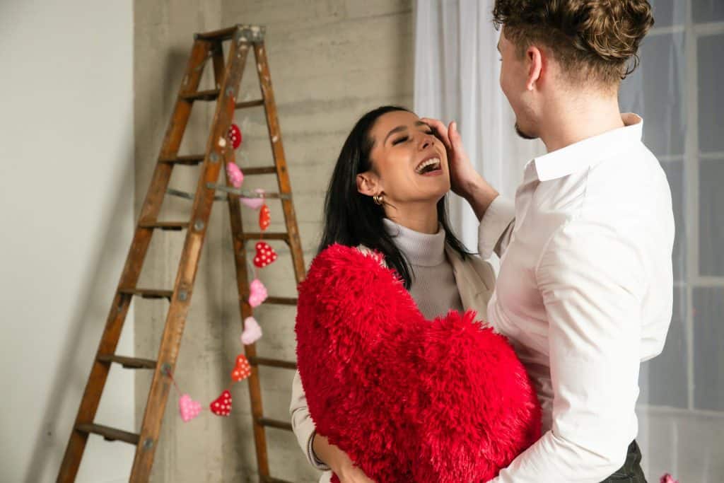 The man is holding a heart-shaped pillow and standing next to the woman.