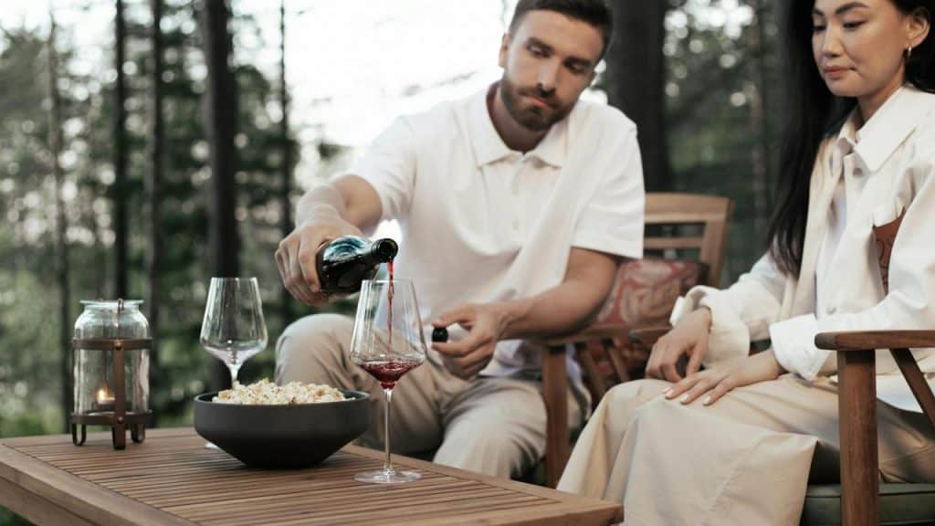A man pouring red wine into a glass while sitting outdoors with a woman.