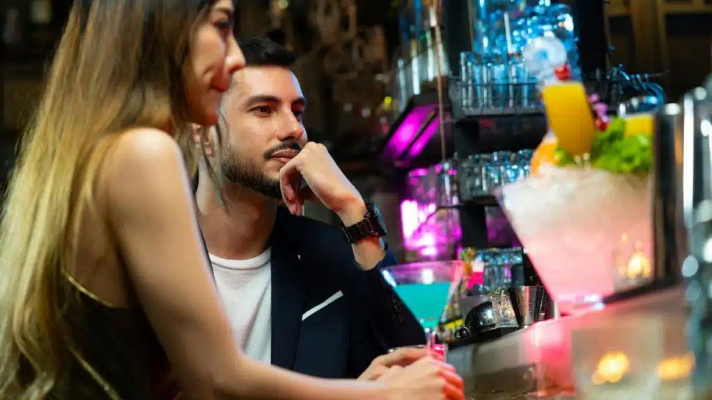 A man and woman sit at a bar with a blue cocktail in the foreground and colorful drinks in the background.