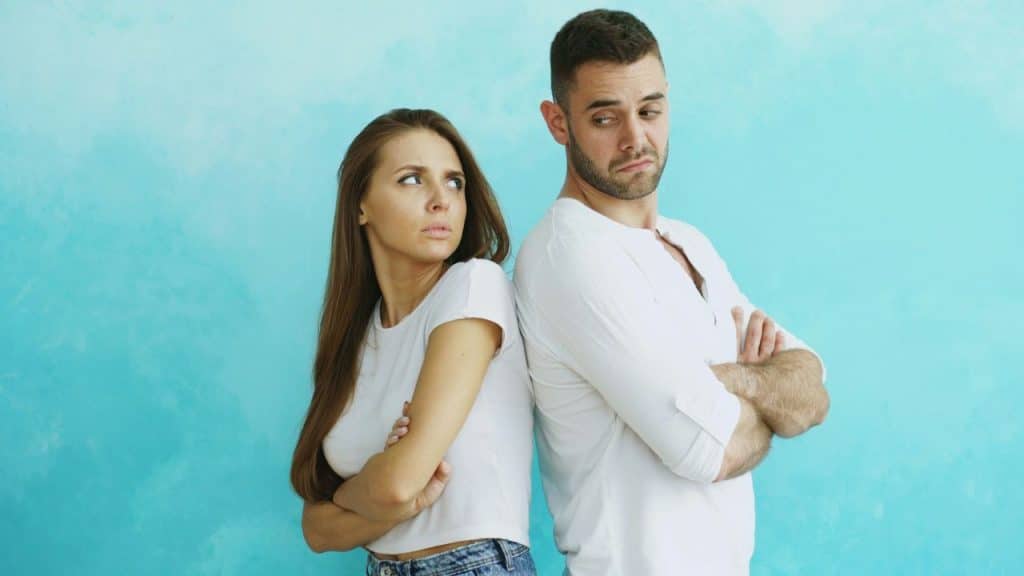 A woman and a man in white shirts stand back-to-back with crossed arms against a blue wall.