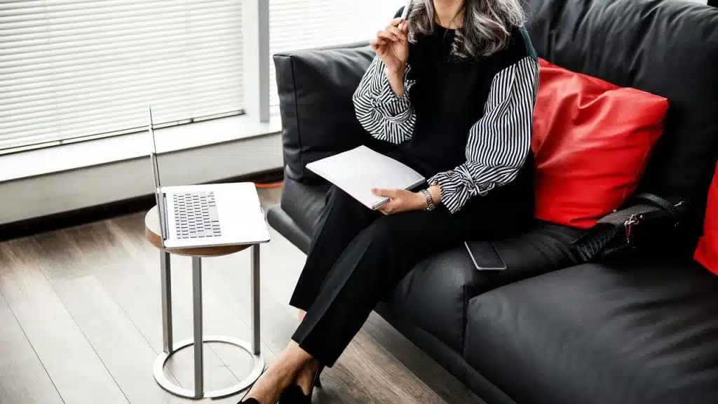 A woman sitting on a couch with a laptop and notebook, appearing focused.