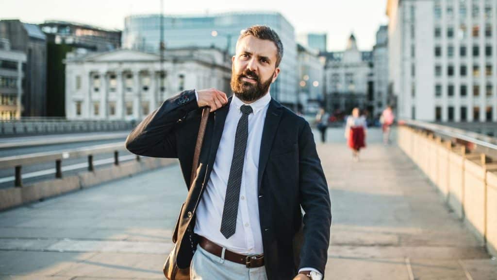 A bearded businessman in a suit is walking across a city bridge during the daytime.