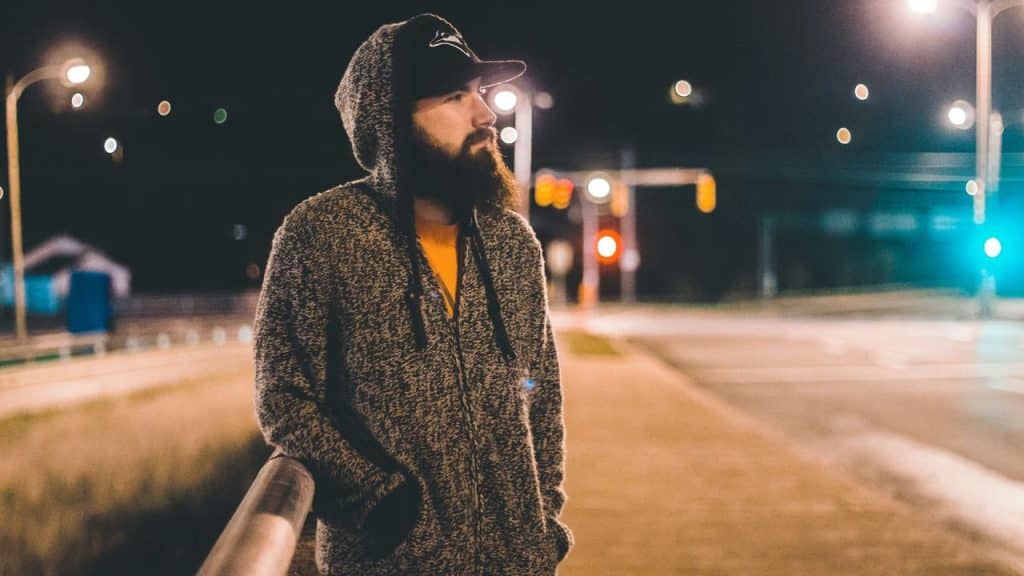 A bearded man in a hooded sweater and cap stands on a bridge at night with streetlights in the background.