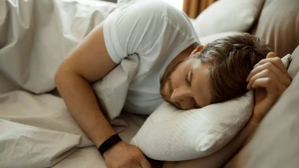A bearded man wearing a white t-shirt and fitness tracker is sleeping on his side in bed.