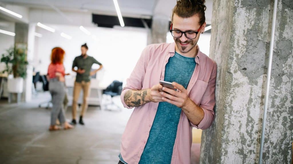A man with glasses and tattoos smiling while using his phone in an office.