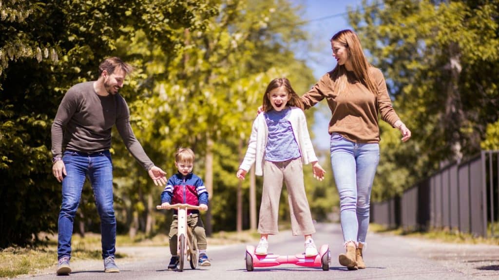 A smiling family walks down a paved road: a man holds a small boy on a balance bike, and a woman walks beside an older girl riding a hoverboard.
