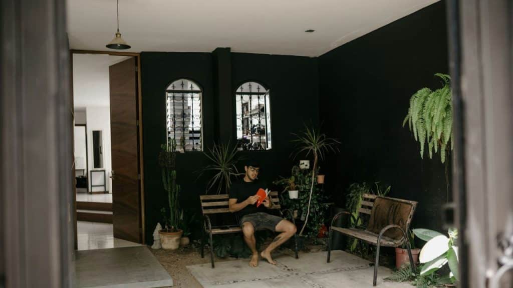 A man sits barefoot, reading a book on a wooden bench in a courtyard with dark walls and many plants.