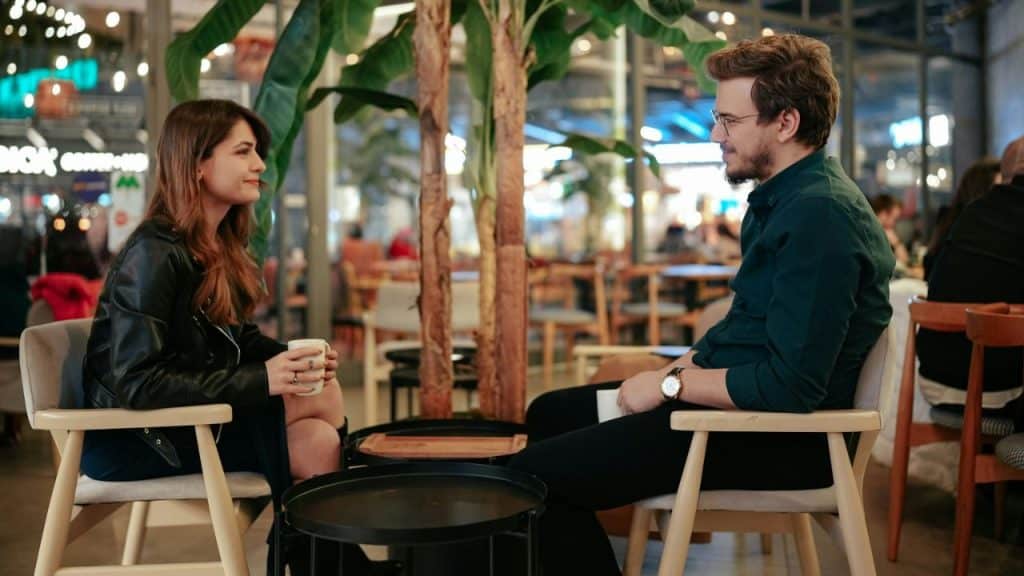 Man and woman sit facing each other in chairs with drinks in an indoor cafe.
