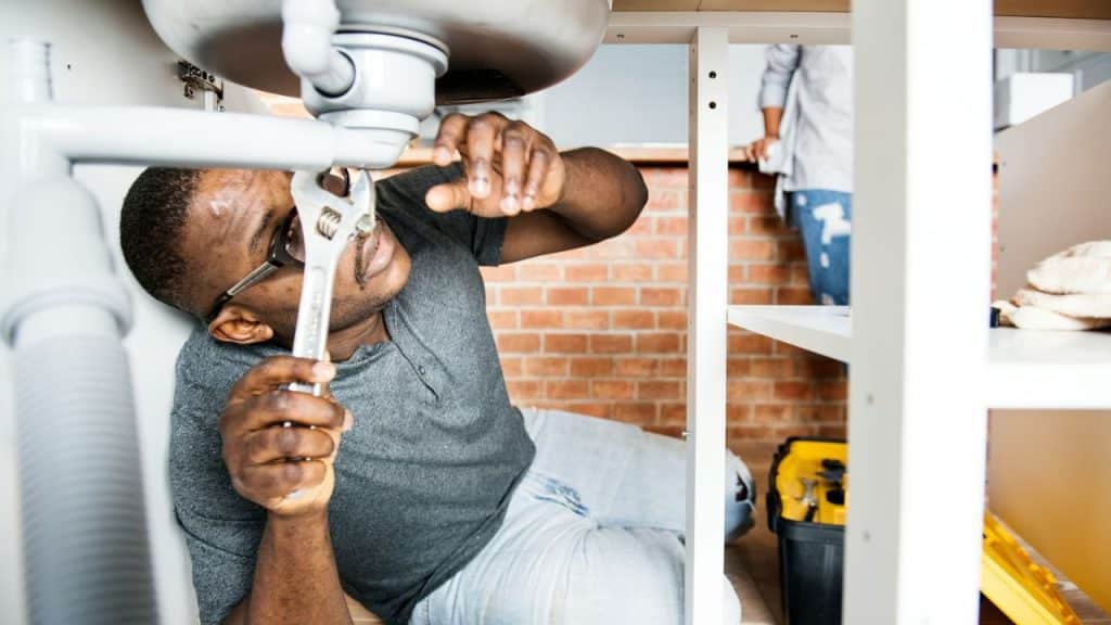 Black man with a wrench fixing a sink pipe while another person stands nearby.