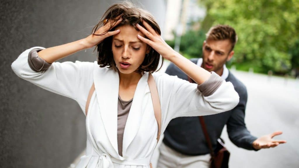 Woman clutching her head in distress while a man gestures behind her outdoors.