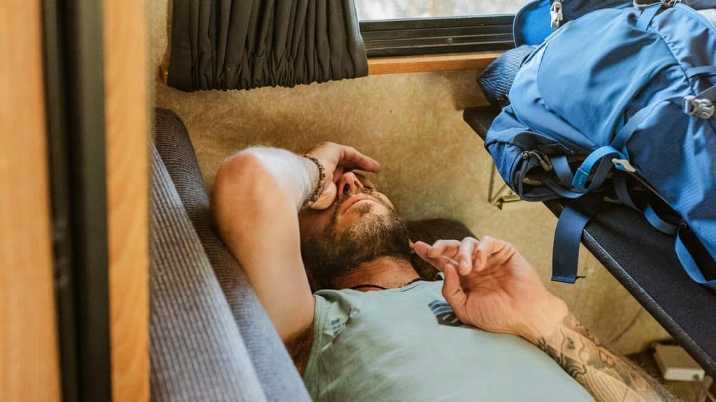 A man lying on a couch resting with his hand on his face beside a blue backpack.