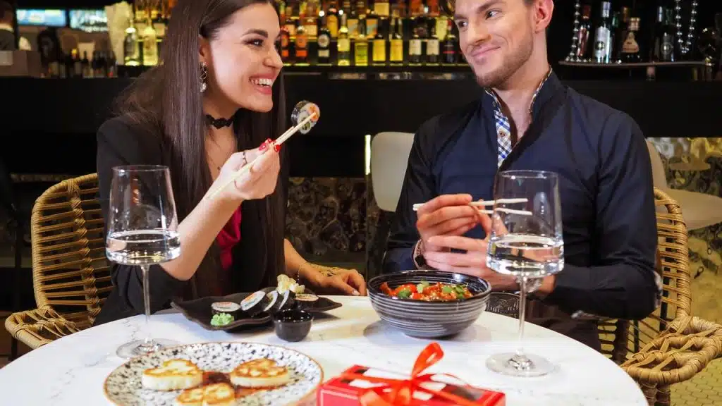 A couple smiling and eating sushi together at a restaurant table.