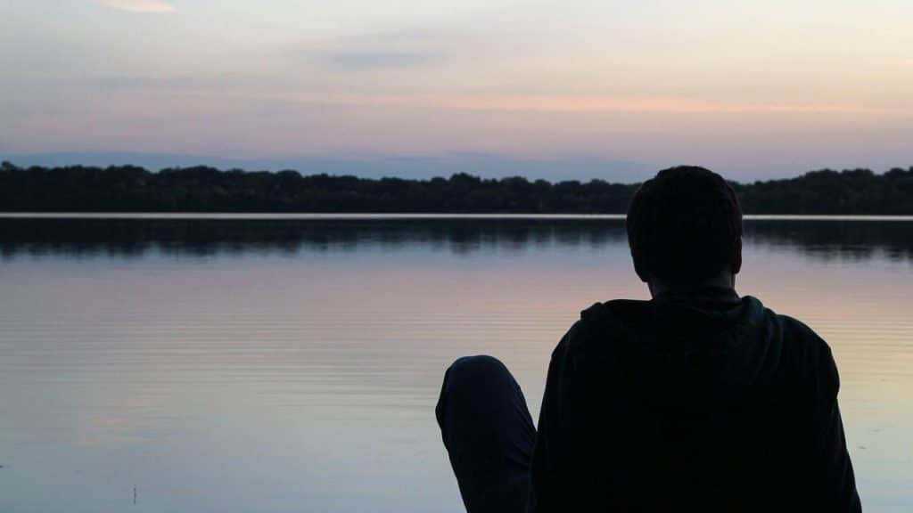 A person sitting by a calm lake at sunset, looking out over the water.