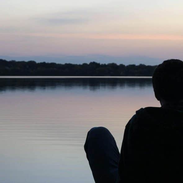 A person sitting by a calm lake at sunset, looking out over the water.