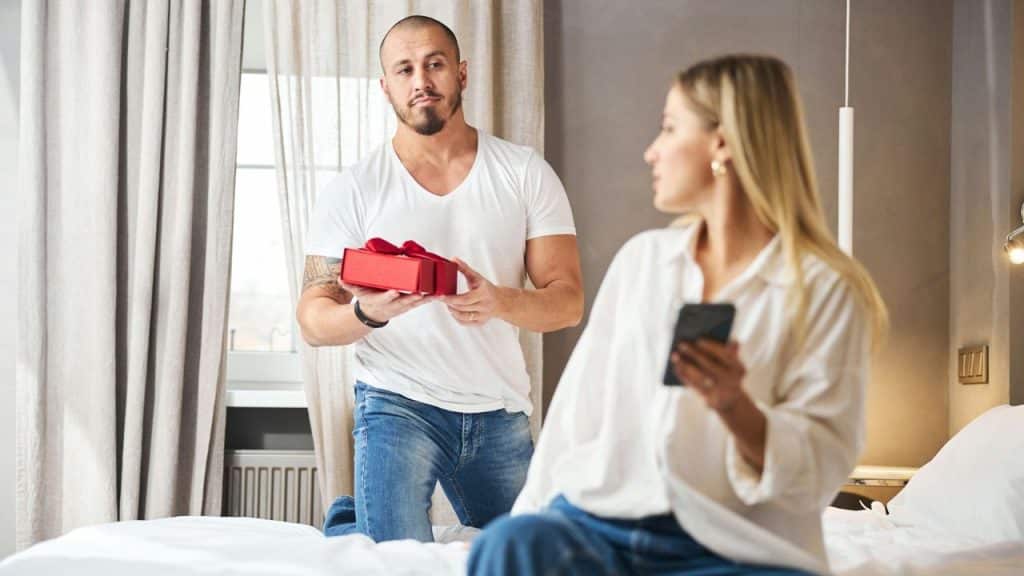 A muscular man kneels on a bed, holding out a red gift box to a blonde woman who is distracted by her smartphone.