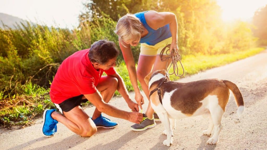 A couple in athletic wear is on a dirt road, with the man kneeling to tie the woman's shoelace next to a beagle.