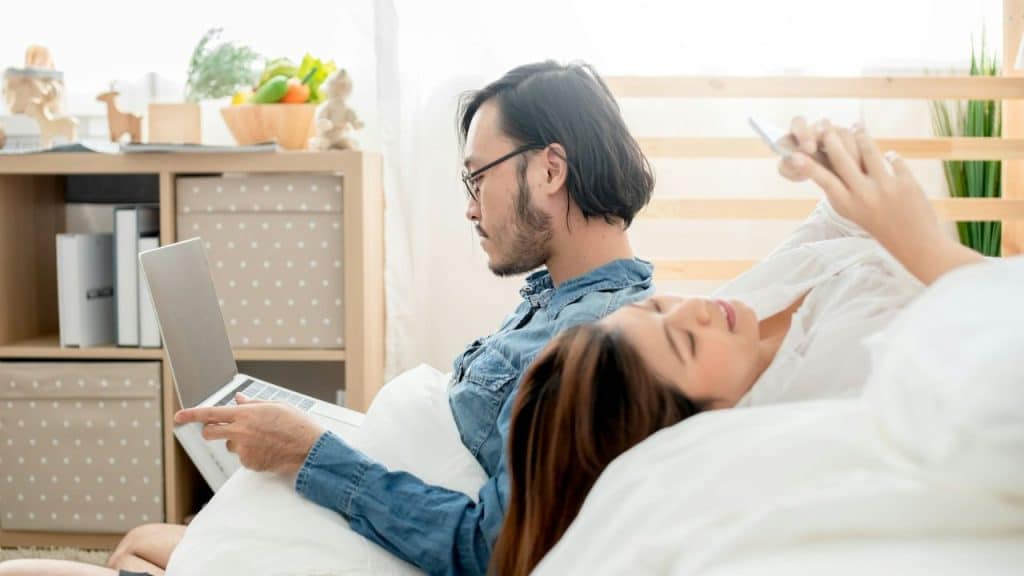 A man works on a laptop while a woman lying next to him uses a smartphone.