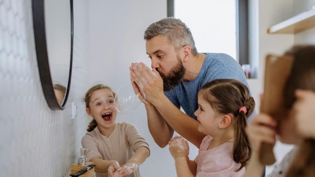 A father having fun blowing bubbles from soap in the bathroom with his little daughters.