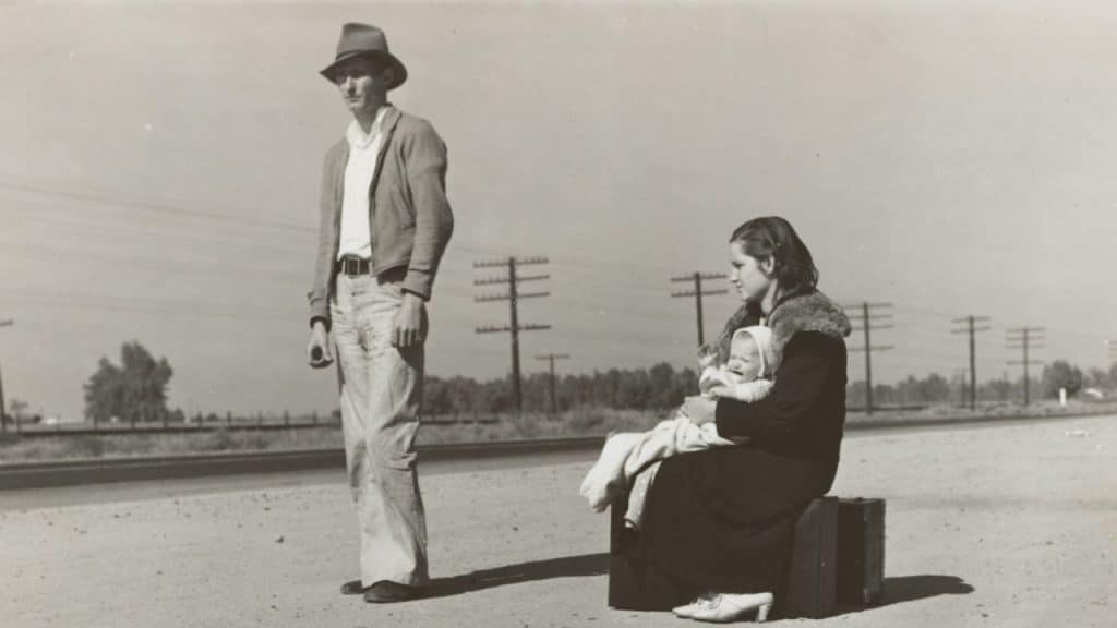 A black and white photo of a man in a hat standing next to a woman seated on a suitcase holding a smiling baby, near railroad tracks.