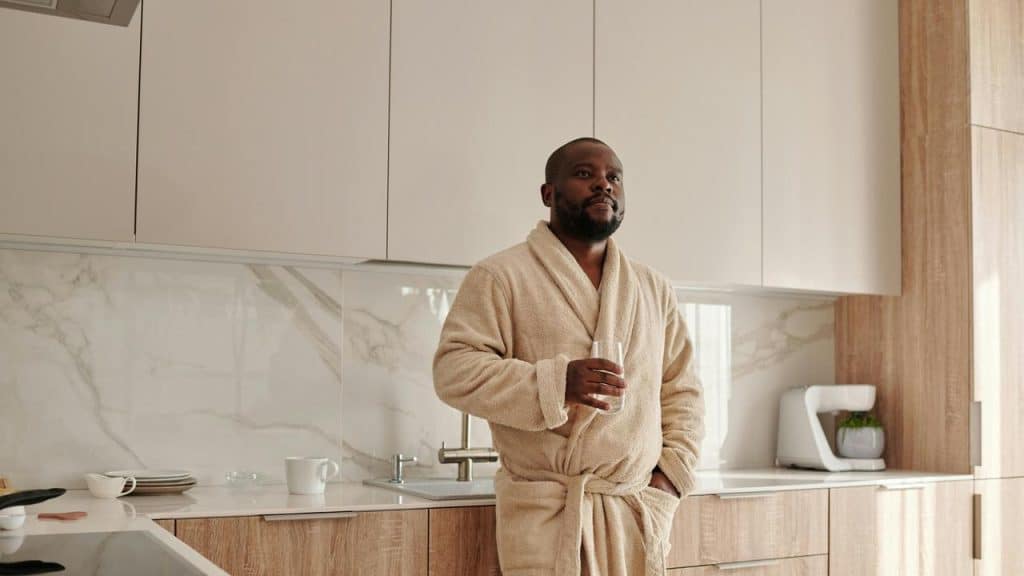 A man in a bathrobe standing in a kitchen holding a glass.