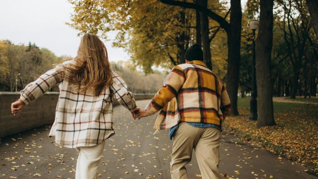 A couple holding hands and running down a tree-lined path covered with autumn leaves.