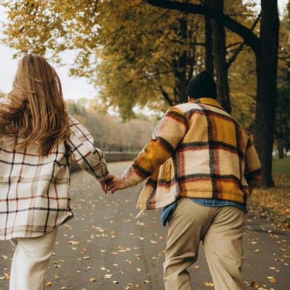 A couple holding hands and running down a tree-lined path covered with autumn leaves.