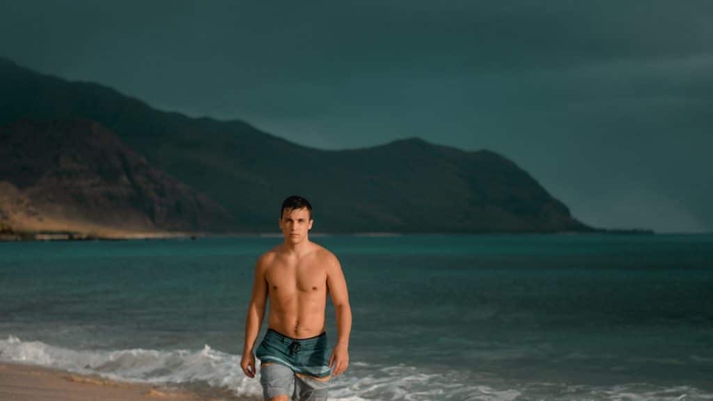 A topless man with blue shorts standing on a seashore during daytime.
