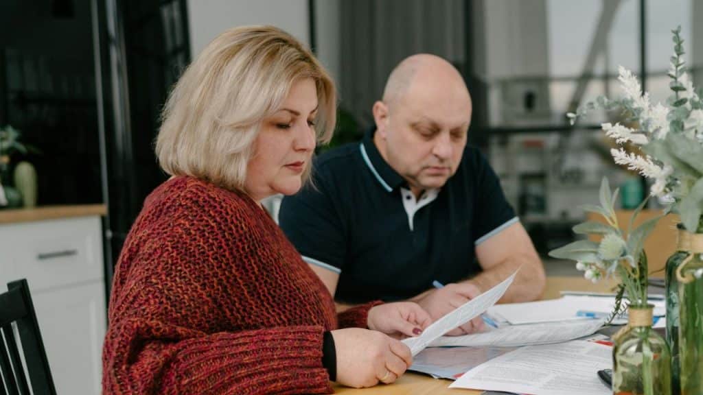 A woman in a red sweater reads a document while a man writes, both seated at a table