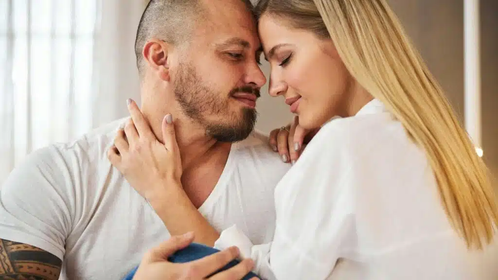 A man and a woman in white shirts are touching foreheads and smiling.
