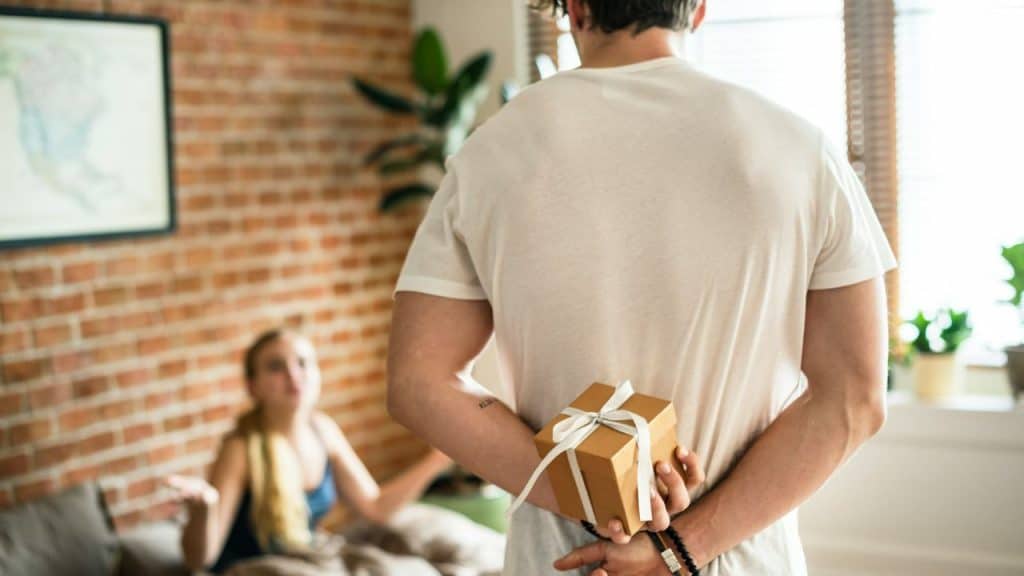 A man, with his back to the camera, holds a small gift behind his back while a woman sits in the blurred background.