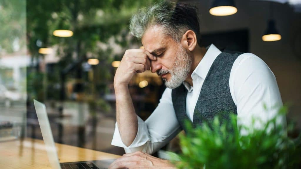 A worried man in a vest and white shirt is sitting with his head in his hand near a laptop.