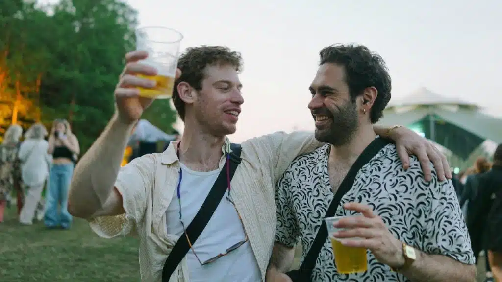Two smiling men are embracing and holding drinks in a crowded outdoor setting.