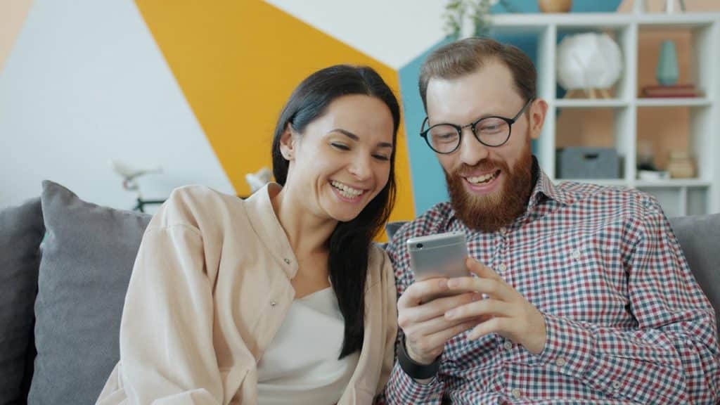 A laughing couple sits on a couch, looking at and smiling at a smartphone held by the bearded man.