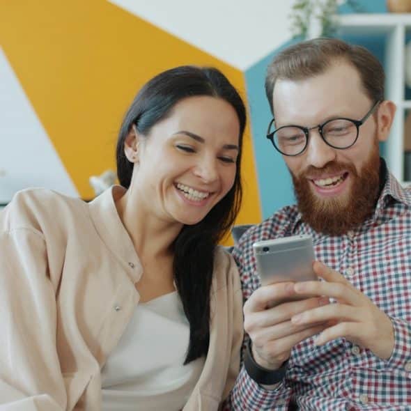 A laughing couple sits on a couch, looking at and smiling at a smartphone held by the bearded man.
