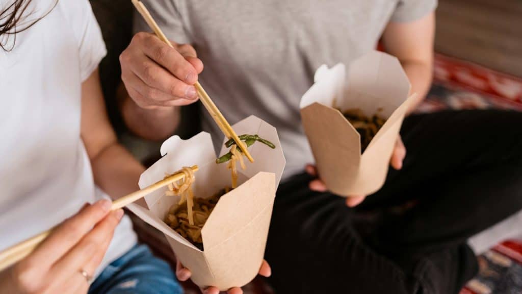 Close-up of a couple eating noodles with chopsticks out of two take-out boxes while sitting down.