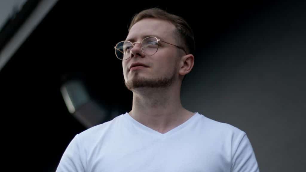 A man with a goatee and round glasses wearing a white V-neck shirt looks up.