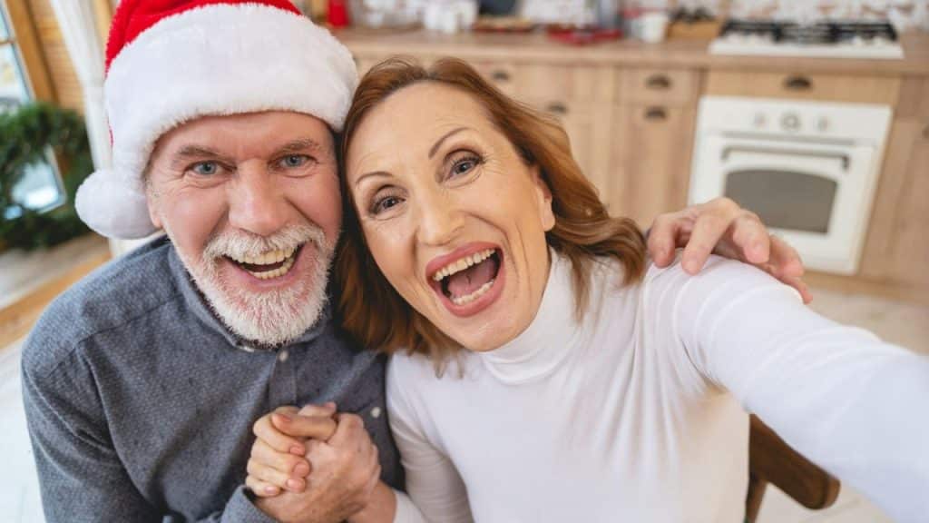 An excited older couple, the man in a Santa hat, holds hands and smiles at the camera.