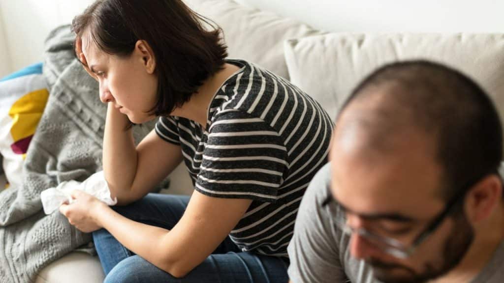 A distressed woman sits on a couch in the foreground; a man's head is out of focus.