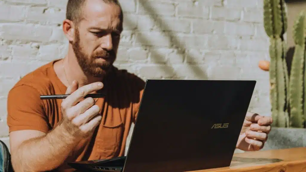 Man with a beard in an orange shirt looking at an ASUS laptop, holding a stylus or pen.