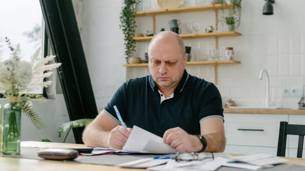 A serious, bald man is reviewing financial documents at a kitchen table.