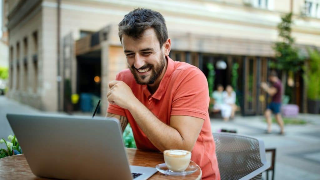 Smiling man at an outdoor cafe using a laptop with a coffee on the table.