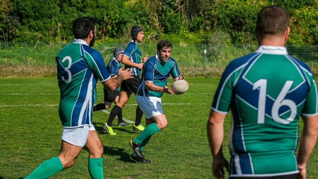 Men in green and blue rugby jerseys are playing with a ball on a sunny field.