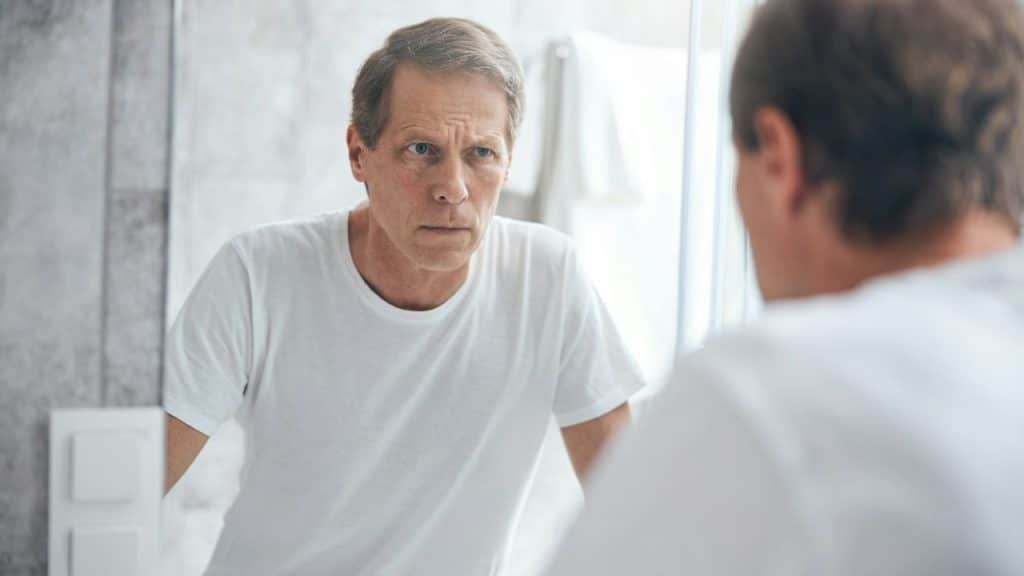 A serious middle-aged man is staring intensely at his reflection in a bathroom mirror.