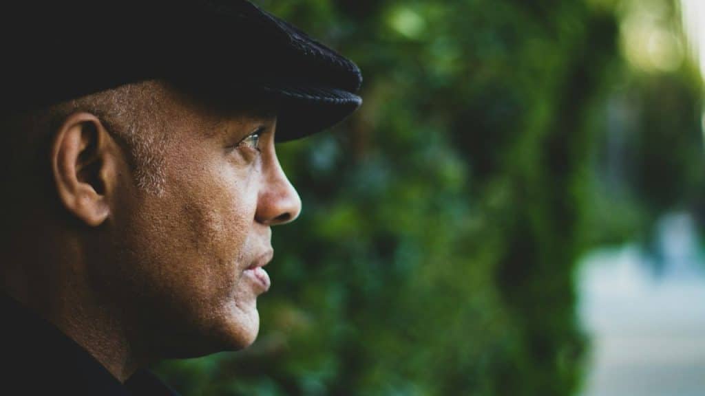 Close-up profile of a man wearing a black flat cap against a blurry green background.