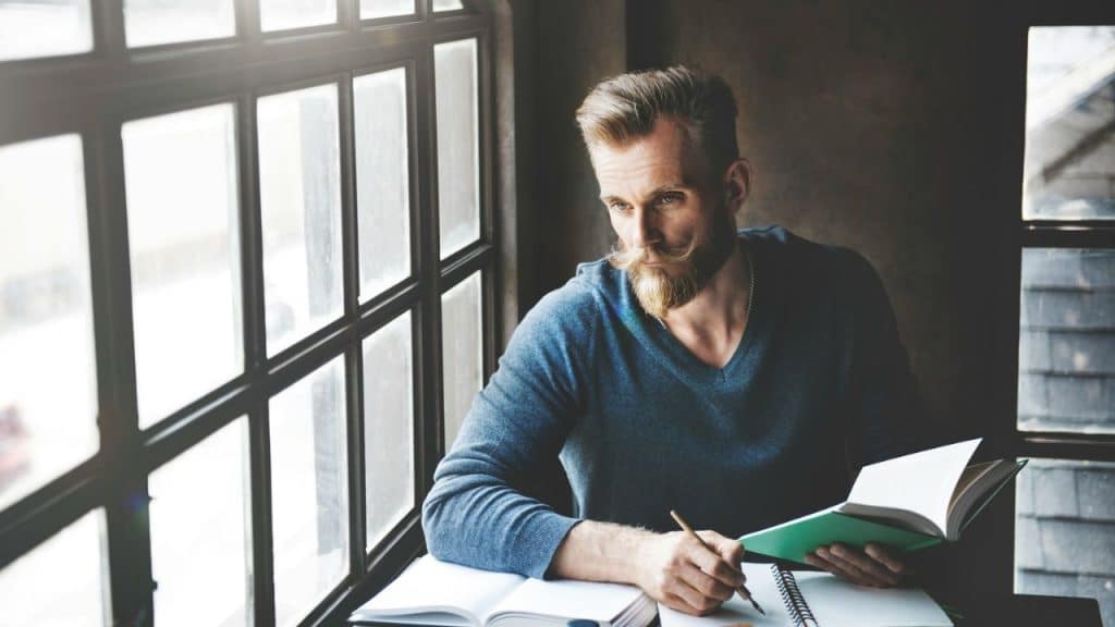 Man with large mustache and beard writes in a notebook by a large, sunny window.
