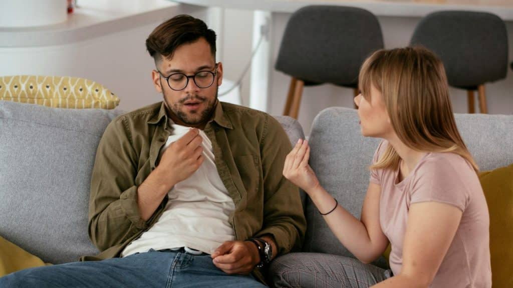 A woman gestures while speaking to a man on a couch with his hand on his chest.