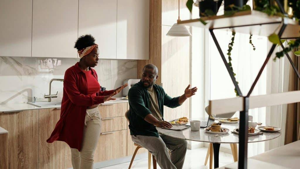 A woman standing and gesturing while talking to a man sitting at a kitchen table.