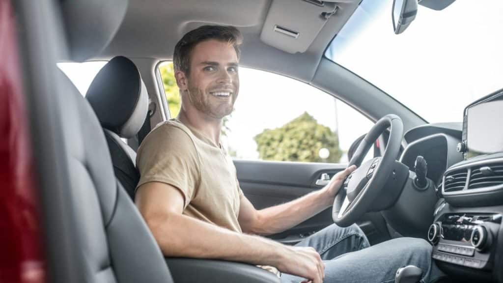 Smiling man in a tan T-shirt and jeans sitting in the driver's seat of a car.