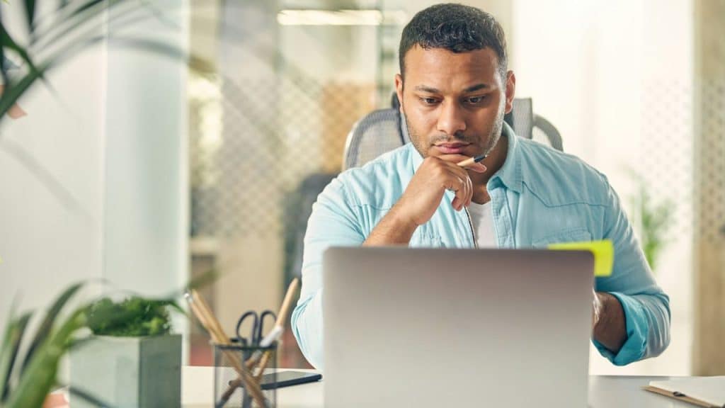 A focused man in a light blue shirt works on a laptop at an office desk.